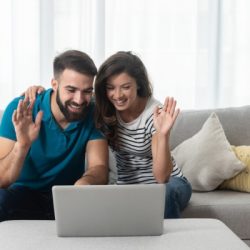 young couple sitting in their living room with laptop waving